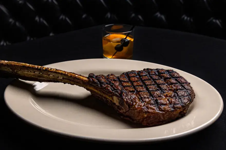 Close-up of a perfectly grilled tomahawk ribeye steak entrée at Prime 47 Carmel, served on a white plate beside a classic Old Fashioned cocktail in a black tufted booth setting