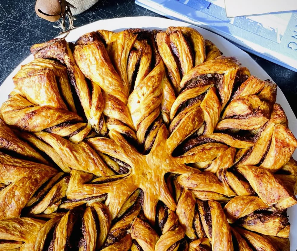 Overhead view of Le Petit Gâteau’s signature Nutella Snowflake Bread, a large round braided pastry with golden-brown flaky layers elegantly arranged in a stunning snowflake pattern, filled with swirls of rich Nutella, presented on a white plate with festive holiday decorations including greenery, pinecones, and a small bell in the background.