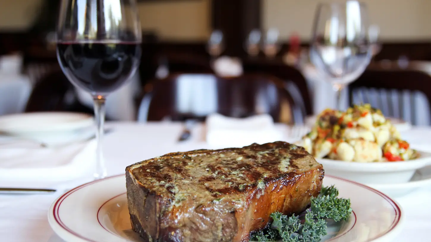 Plated dry-aged steak served alongside a glass of red wine on a white table setting at P.S. Steak in Minneapolis, Minnesota