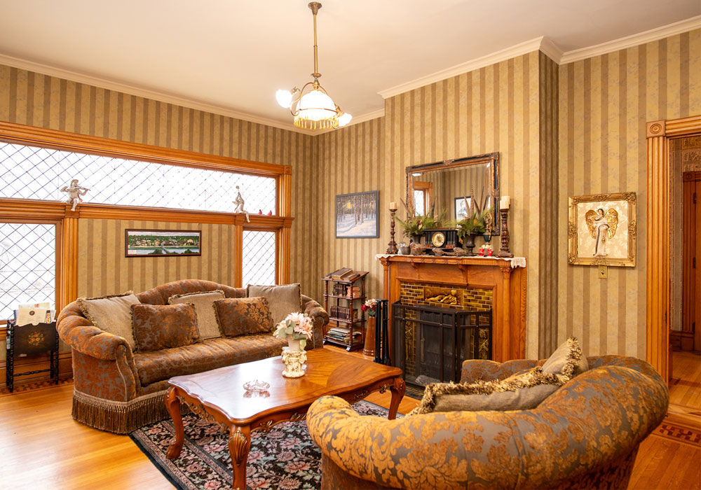 Elegant common parlor interior at Aurora Staples Inn featuring striped wallpaper, a large patterned sofa with pillows, a wooden coffee table, a second curved sofa in the foreground, a brick fireplace with wooden mantel and mirror above it, large windows with diamond-patterned glass, hardwood floors with a decorative rug, and a chandelier overhead.