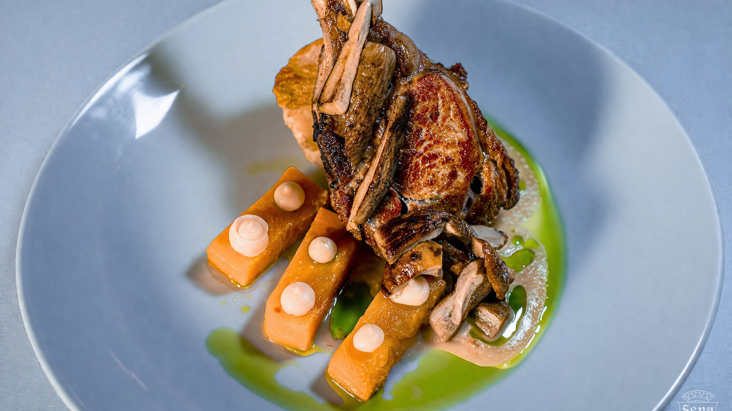 Elegantly plated grilled lamb chop with long frenched bone standing upright, accompanied by glazed orange vegetable batons topped with cream dollops, sautéed mushrooms, and artistic swirls of vibrant green and beige sauces on a white plate at La Famiglia Ristorante.