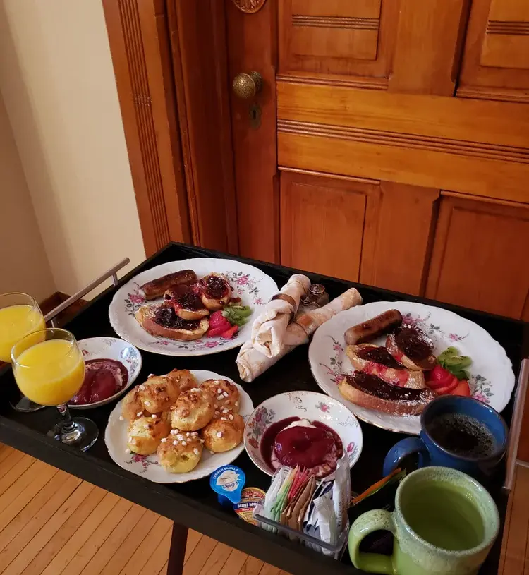 Generous black rolling breakfast tray at Ann Bean Mansion Bed & Breakfast loaded with two white floral plates of sausage links, French toast or bagels topped with red jam and strawberries, a plate of golden pastries, glasses of fresh orange juice, a blue coffee mug, a green ceramic tea mug, small bowls of preserves, rolled napkins with silverware, and assorted condiments, all set on polished hardwood flooring in front of a warm wooden paneled door.