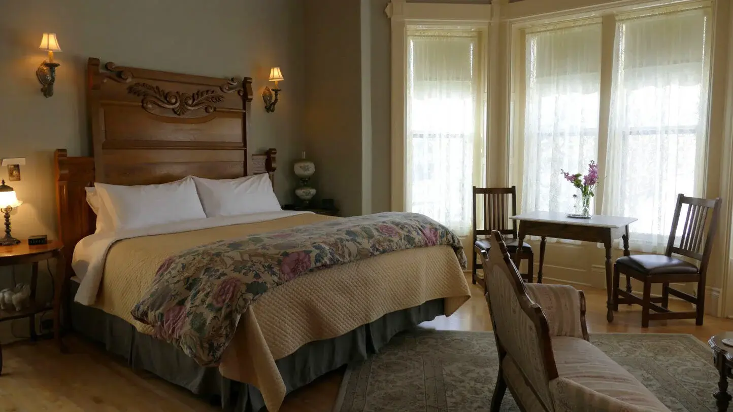 Spacious and refined guest bedroom interior at Ann Bean Mansion Bed & Breakfast featuring a grand dark wood bed with an intricately carved ornate headboard, white pillows, and a floral-patterned quilt in soft beige and pink tones, flanked by matching wall sconces, large bright windows with sheer curtains flooding the room with natural light, a small wooden table and chairs by the window with a vase of fresh purple flowers, a patterned area rug on hardwood floors, and an upholstered chaise or chair in the foreground.