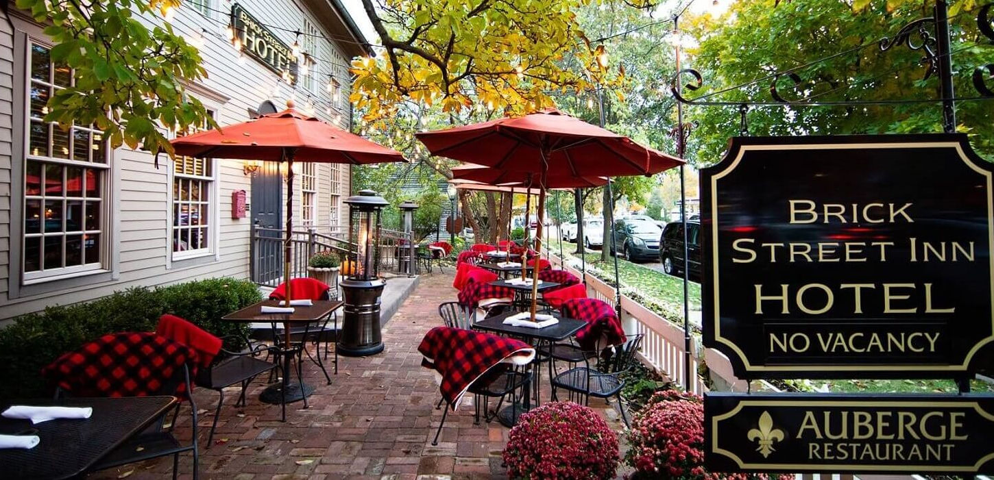 Wide view of the inviting brick-paved patio at Brick Street Inn in Zionsville, Indiana, showing multiple tables with red umbrellas, chairs draped in red-and-black checkered blankets, autumn foliage and string lights overhead, flowerbeds in the foreground, and prominent black signs for Brick Street Inn Hotel and Auberge Restaurant.  