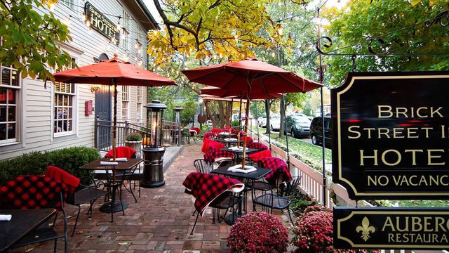 Wide view of the inviting brick-paved patio at Brick Street Inn in Zionsville, Indiana, showing multiple tables with red umbrellas, chairs draped in red-and-black checkered blankets, autumn foliage and string lights overhead, flowerbeds in the foreground, and prominent black signs for Brick Street Inn Hotel and Auberge Restaurant.  