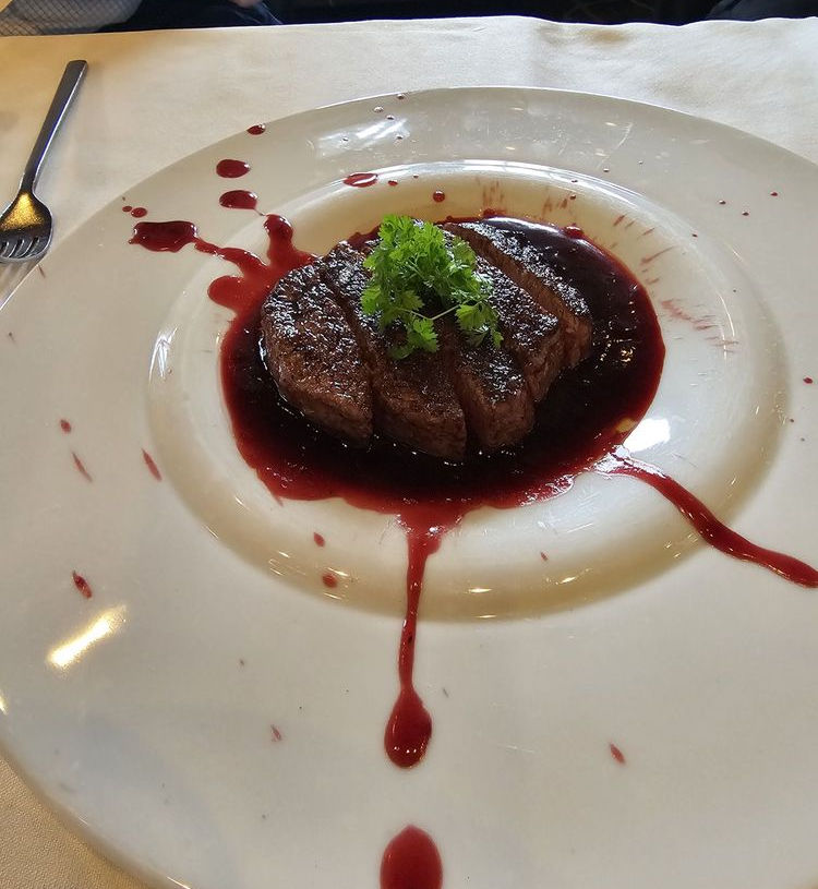 Close-up of an elegantly plated sliced filet mignon at Anthony’s Chophouse in Carmel, Indiana, featuring a dark seared crust and juicy medium-rare pink interior, nestled in a rich red wine reduction sauce with artistic drips and splatters across a pristine white plate, topped with a fresh parsley sprig
