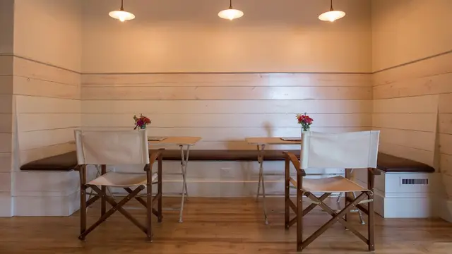 Cheerful and cozy seating/breakfast area at Hotel Broad Ripple featuring light-colored horizontal shiplap walls, built-in benches with dark cushions, two wooden director-style chairs with white canvas fabric, small folding tables with colorful flower vases, three pendant lights hanging from the ceiling, and warm hardwood floors.  