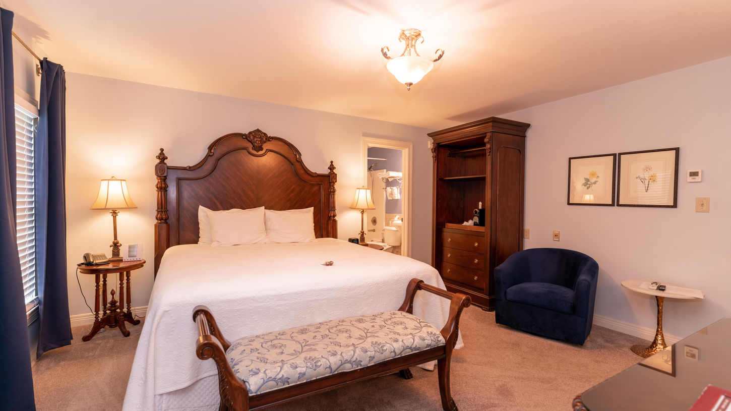 Spacious, warmly lit guest room at Brick Street Inn in Zionsville, Indiana, featuring a large king bed with an elaborate carved dark wood headboard and footboard, crisp white quilted bedding and pillows, a decorative wooden bench with floral-patterned upholstery at the foot of the bed, a tall wooden armoire/wardrobe on the right, a cozy blue upholstered armchair, blue curtains framing a window, framed floral artwork on the walls, and soft ceiling lighting creating an inviting, luxurious atmosphere.  