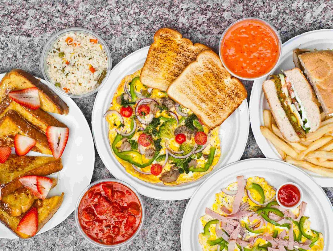 Overhead view of a generous brunch platter selection at Hen House Eatery in Minneapolis including French toast with strawberries, veggie omelette with toast, breakfast sandwich with fries, and side sauces on a marble table.