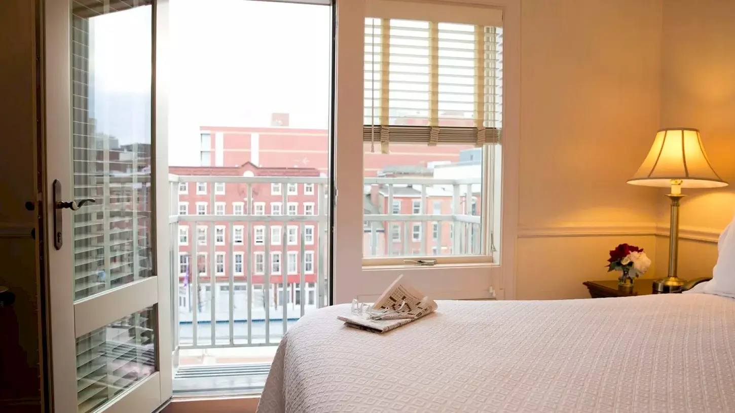Bright and inviting king guest room at Penn’s View Hotel featuring a large bed with white quilted linens and a folded newspaper, an open glass door leading to a private balcony overlooking historic red-brick buildings in Philadelphia, a window with blinds, and a warm bedside lamp with fresh flowers.