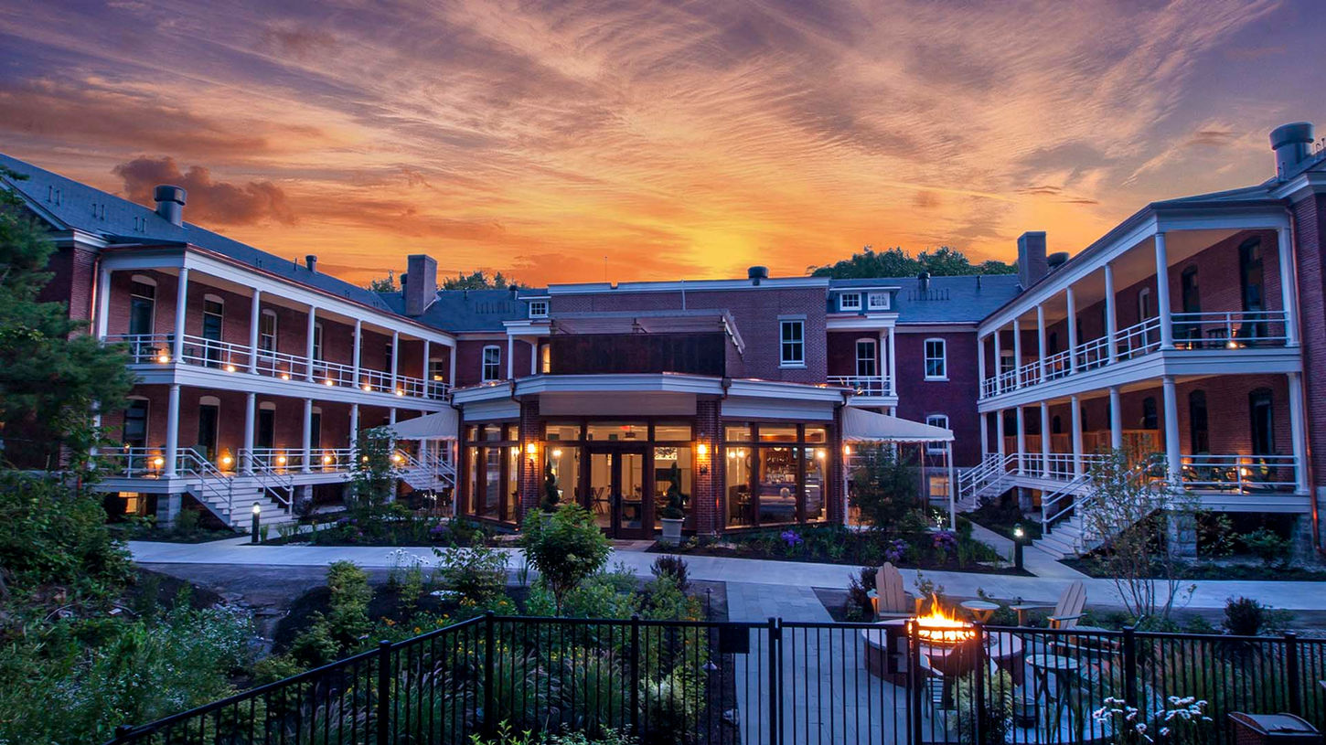 Stunning sunset view of the historic red-brick Inn at Diamond Cove buildings on Great Diamond Island with glowing porches, manicured gardens, and fire pit overlooking Casco Bay.