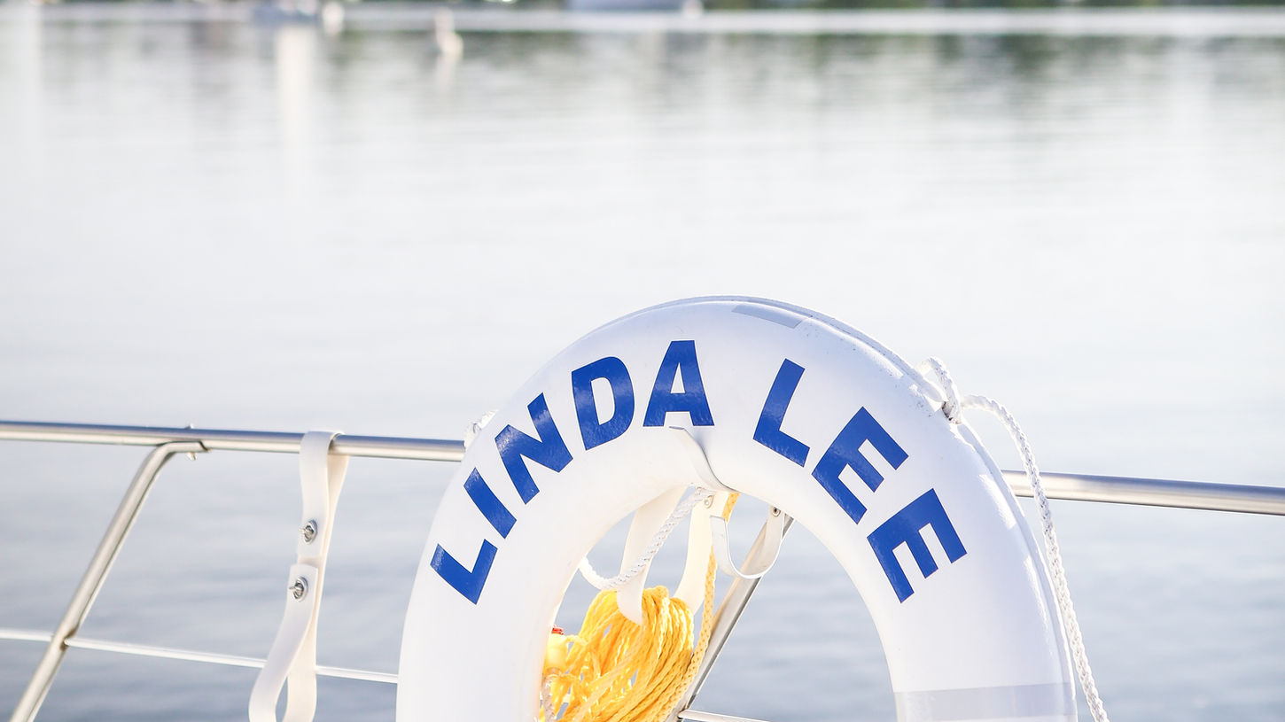 Close-up of the white life ring with bold blue lettering “LINDA LEE” and “Excelsior, MN” prominently displayed on the railing of the luxury yacht, with calm Lake Minnetonka waters and scenic green shoreline in the background on a bright sunny day.