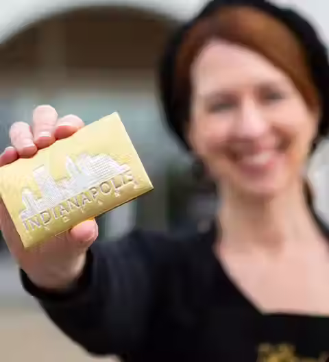 A smiling woman with reddish-brown hair and a black beret proudly holds up a luxurious gold chocolate bar embossed with a detailed Indianapolis skyline and the word “INDIANAPOLIS,” showcasing DeBrand Fine Chocolates’ artisan craftsmanship and city-inspired design in sharp, vibrant detail.