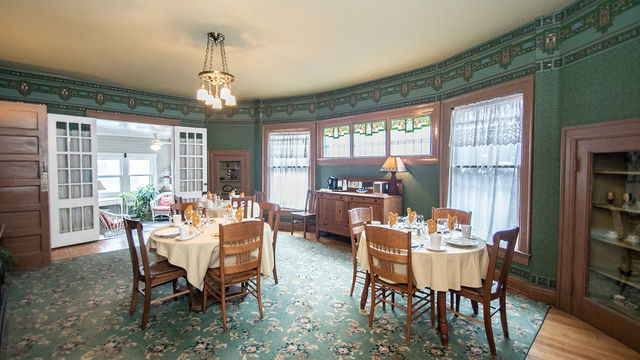 Spacious and inviting dining room at Looking Glass Inn featuring lush green floral-patterned carpet and coordinating wallpaper border with decorative molding, multiple round wooden tables elegantly set with white tablecloths, china, glassware, napkins, and fresh flowers, surrounded by classic wooden dining chairs, a brass chandelier overhead, large windows with sheer curtains and stained-glass transoms, a wooden buffet sideboard, and open doorways revealing adjacent historic rooms.  