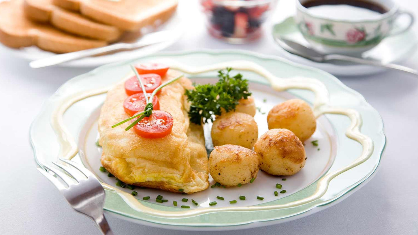 Elegant plated breakfast at The Inn at Bowman’s Hill featuring a golden omelette garnished with sliced cherry tomatoes on a chive tie and fresh parsley, accompanied by small roasted potatoes and chives on a light green scalloped plate. In the softly blurred background are slices of toast, a bowl of mixed berries, and a cup of coffee with saucer.