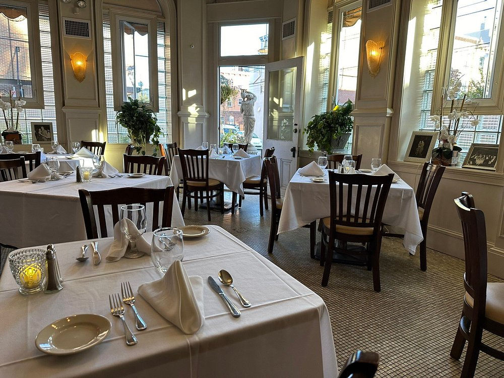 Brightly lit daytime view of the elegant dining room at Dante & Luigi’s restaurant in Philadelphia, featuring white tablecloths, dark wood chairs with formal place settings, large windows flooding the space with natural light, potted plants, wall sconces, and a view through the open door to a statue outside.