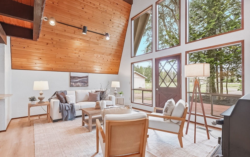 Cozy living room with wooden ceiling, large windows showing trees, cream sofa with cushions, a light toned coffee table, and two cream accent chairs. Soft lighting from lamps and serene decor.