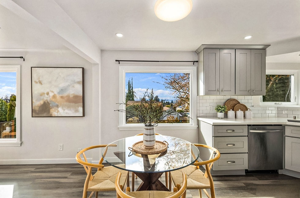 Bright kitchen-dining area with a glass table and wooden chairs. A vase with branches sits on the table. Large windows offer a view outside.