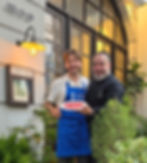 Jose Sol and Chef Pepe Dasi smiling outside Luma restaurant, holding a plate of Jamón Ibérico, surrounded by green plants and warm evening light
