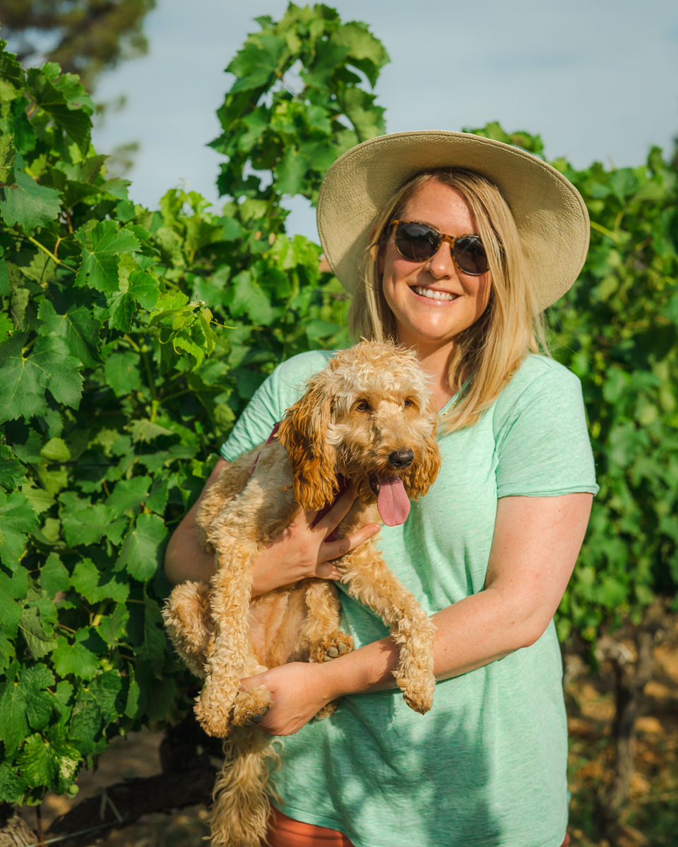 a blonde woman in a green shirt in a vineyard with a cockapoo puppy