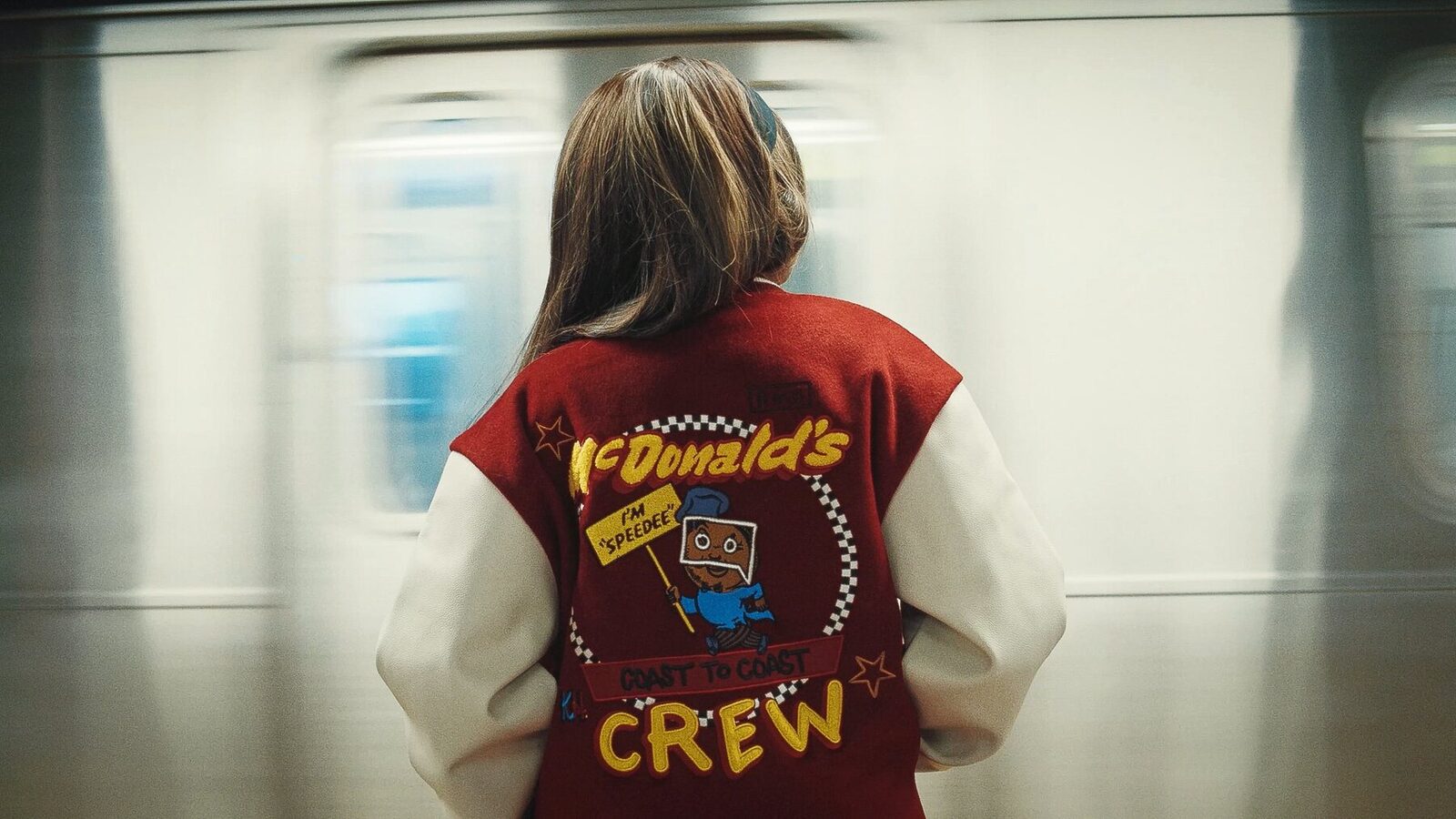 A kid in a red McDonald's crew jacket watches a train blur past on a subway platform