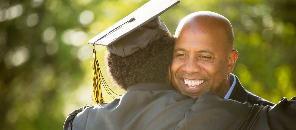 A father gives his college son, who as just graduated, a hug. The son is wearing a graduation cap and gown.