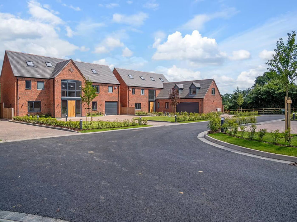 A row of three modern, red-brick houses on a curved street under a blue sky with clouds. The scene is calm and picturesque, with neatly trimmed lawns.