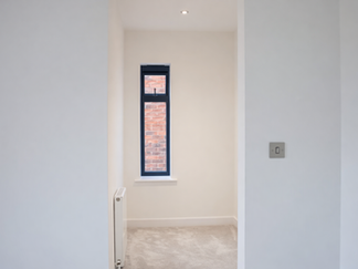 Small neutral bedroom with cream carpet, narrow black-framed window, white walls, and a radiator beneath the window.