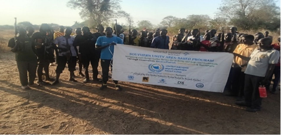 Peace committee members and armed youth hold a banner for the Southern Unity Area-Based Program outdoors after mediation dialogue. Dry landscape background.
