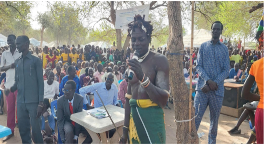 Spear master speaks into a microphone at an outdoor gathering, surrounded by a crowd under trees.