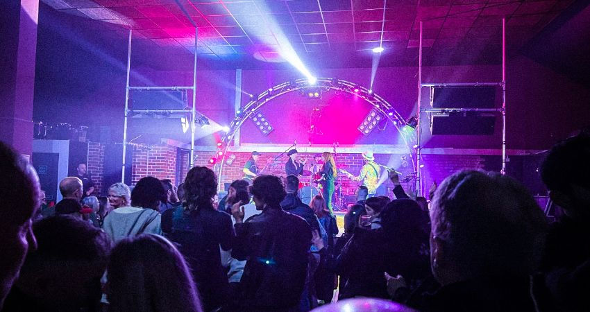 Crowd watches a band perform on stage with vibrant pink and blue lights in a club setting, creating a lively and energetic atmosphere.