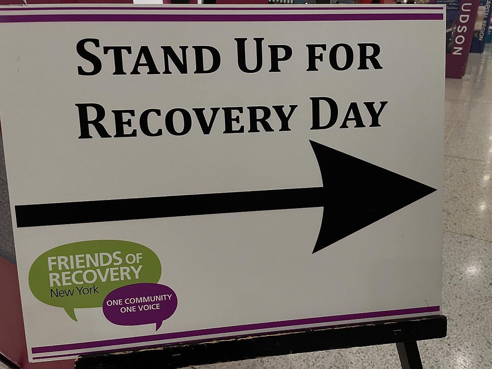 Sign with black text "STAND UP FOR RECOVERY DAY" and an arrow. Logo reads "FRIENDS OF RECOVERY New York." Indoor setting with shiny floor.