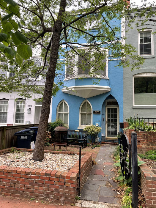 A sidewalk view with boxed brick raised bed and flagstone/brick front patio area. The iron gate has a circular design. (D. Vogt, May 2022)