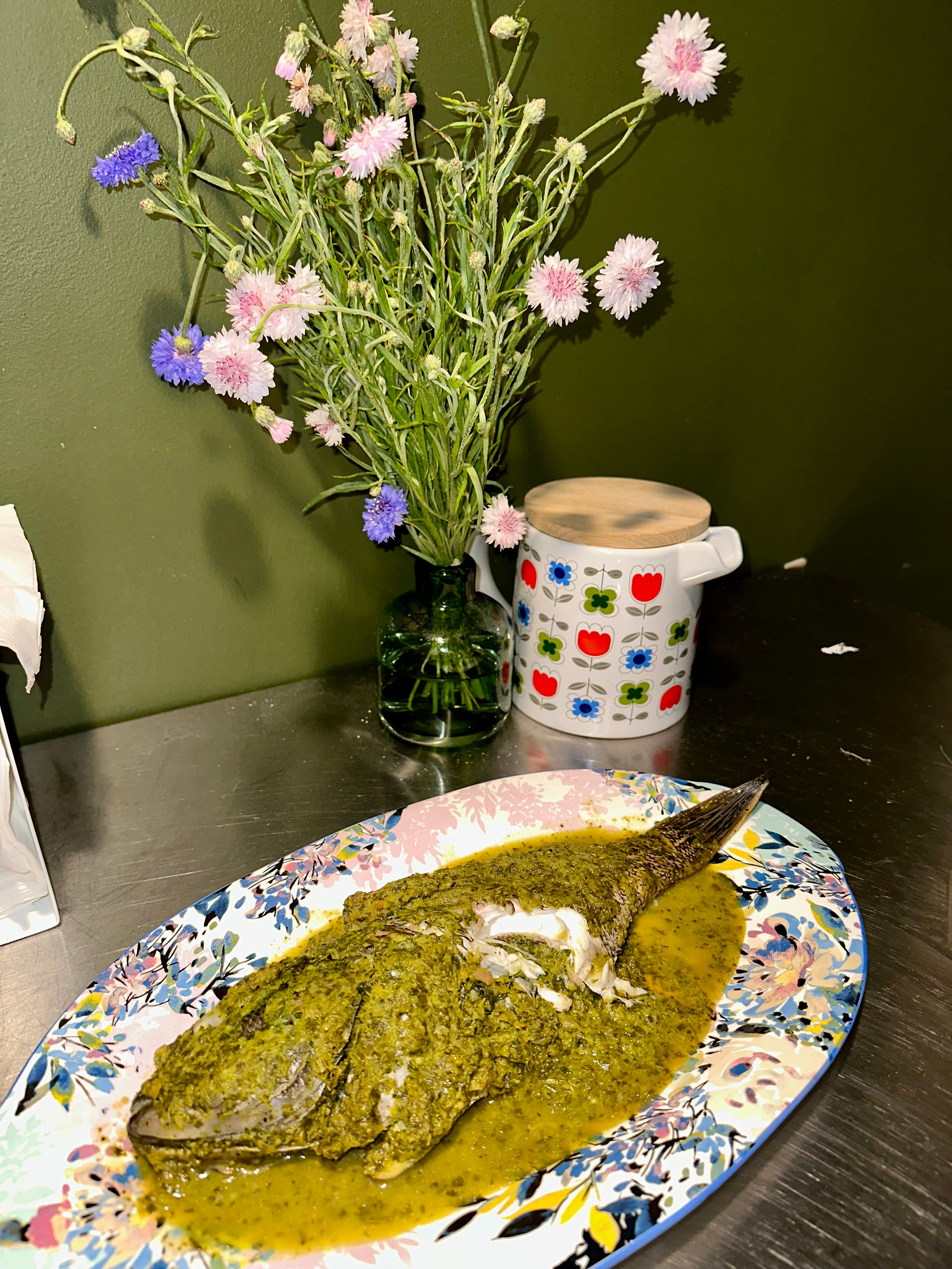 a baked seabass covered in an herb and vegetable mixture sits on a blue and purple floral plate. a bouquet of flowers is in the background