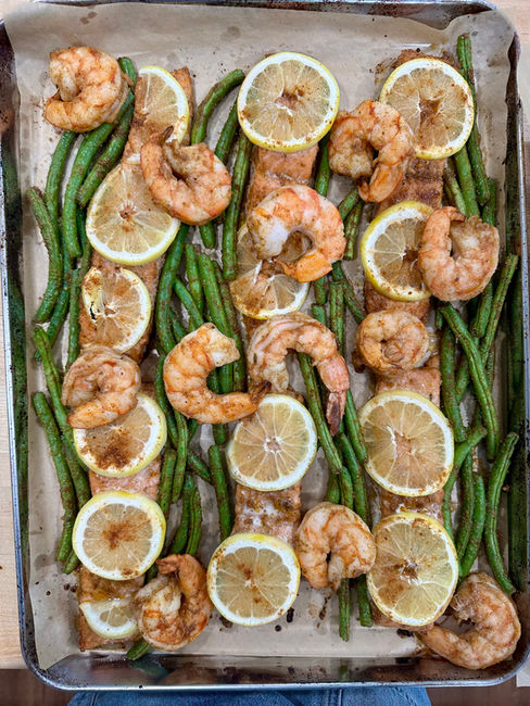 A sheet tray lined with parchment topped with baked shrimp, salmon, green beans, and lemon slices