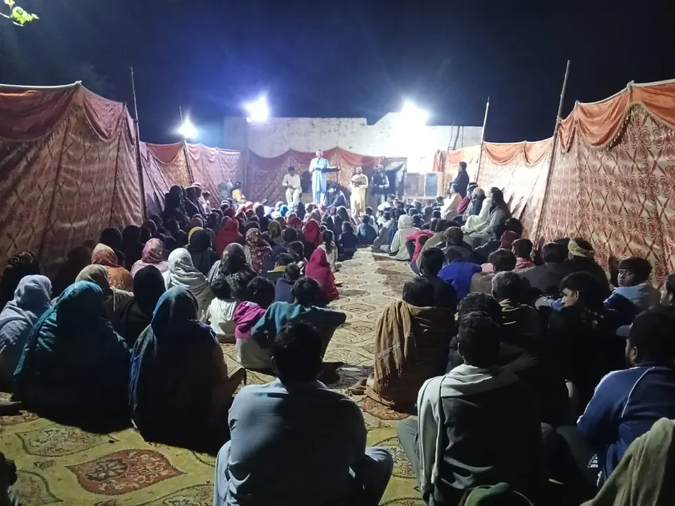 A crowd gathers in a tented area at night, facing a small stage with speakers. The scene is lit by bright lights, creating a focused atmosphere.