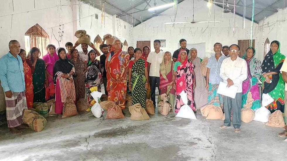 Group of people in colorful clothes standing in a room with bags of goods on the floor, suggesting a distribution event. Neutral expressions.