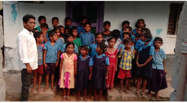 A group of children in colorful school uniforms stands outside a building with a teacher. The mood is neutral; background is plain.