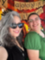 Two women smiling in front of a festive display with burlap flags that read “Goodies Always.” One wears black cat-eye sunglasses and has long gray hair; the other wears clear glasses and a green top.