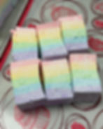 Six pastel rainbow marshmallows arranged on a patterned surface, each featuring soft, even layers of pink, yellow, green, blue, and purple.