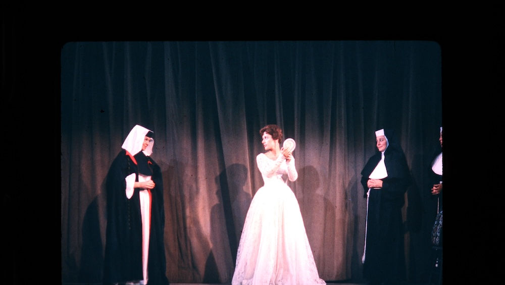 Woman in white gown performs on stage, flanked by two nuns.