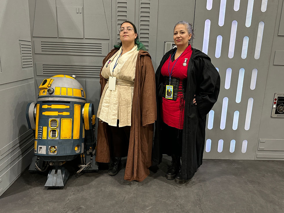 Two women in Star Wars cosplay pose confidently in front of a sci-fi backdrop. One wears Jedi robes with a brown cloak, and the other wears a red and black outfit with a long black coat. A yellow and blue astromech droid is partially visible beside them.