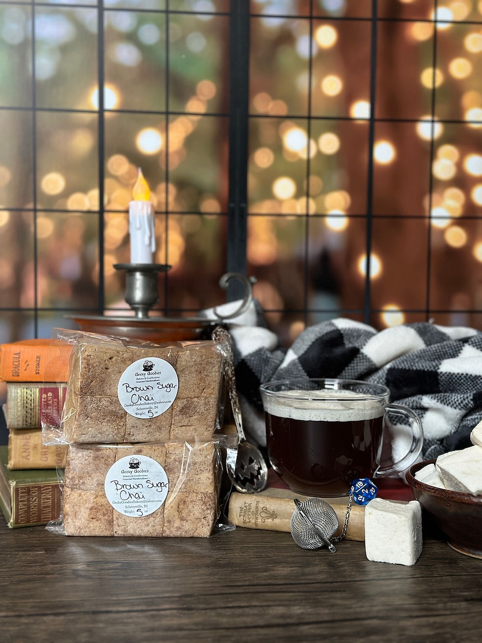 A cozy, autumn-themed setup featuring two wrapped "Brown Sugar Chai" marshmallow packs from Geeky Goodies, placed on top of vintage books. A clear mug of dark tea, a black-and-white plaid blanket, and a candle add warmth to the scene. A tea infuser, a blue 20-sided die, and a marshmallow are arranged nearby, all set against a softly lit backdrop of bokeh lights through a windowpane.