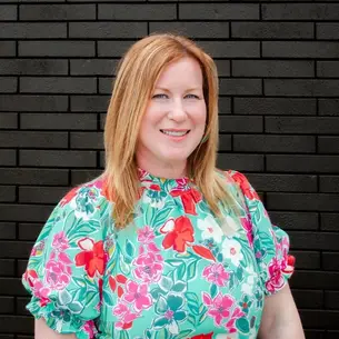 Photo over Melissa Griffin. Owner of Indigo Ink Interiors from valpo, but serving Indiana and Southern Michigan. She has red hair and is wearing a brightly colored mint shirt with red white and pink florals and is standing behind a black brick wall