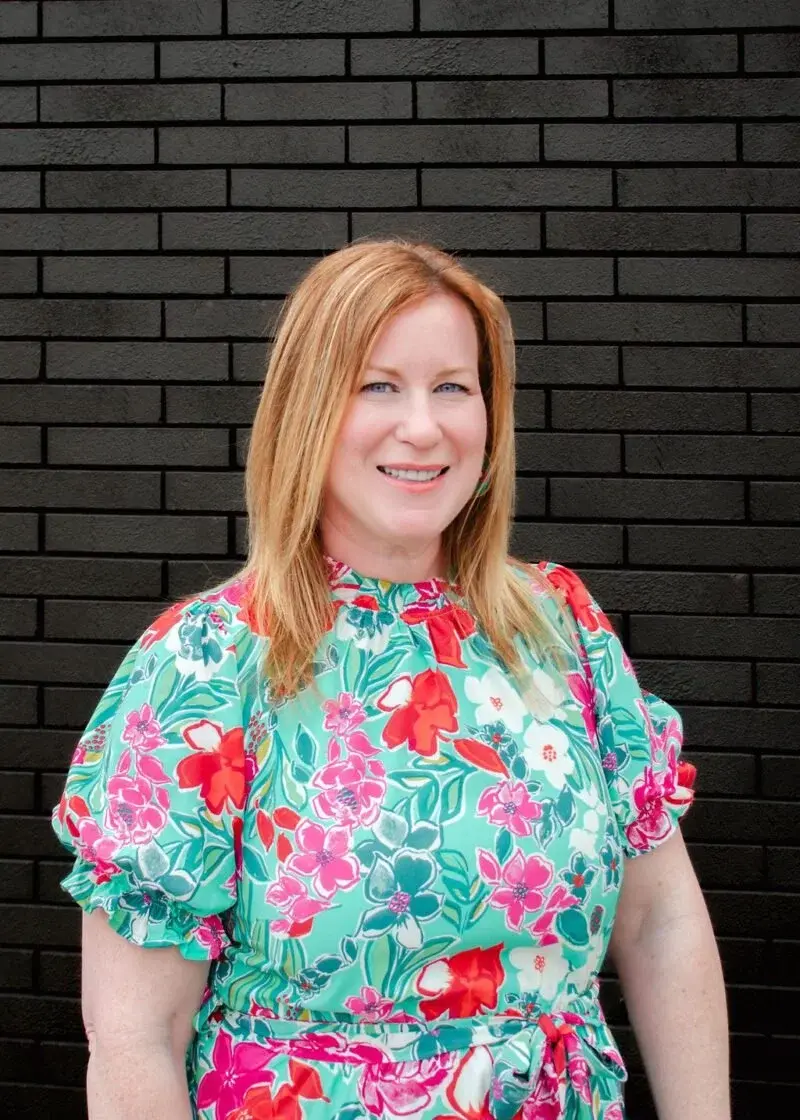 Photo over Melissa Griffin. Owner of Indigo Ink Interiors from valpo, but serving Indiana and Southern Michigan. She has red hair and is wearing a brightly colored mint shirt with red white and pink florals and is standing behind a black brick wall