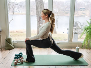 Woman lunges on a yoga mat indoors, facing large windows with a lake view. Dumbbells nearby. Calm and focused mood.