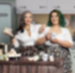 Two women stand smiling in a retro-inspired kitchen, wearing white blouses and geeky-themed skirts—one patterned with chocolate truffles, the other with potions and scrolls. One holds a mug labeled "GG" while the other presents a package of marshmallows from “Geeky Goodies.” A wooden counter in front of them displays colorful marshmallows, polyhedral dice, and jars, blending baking charm with nerdy whimsy.