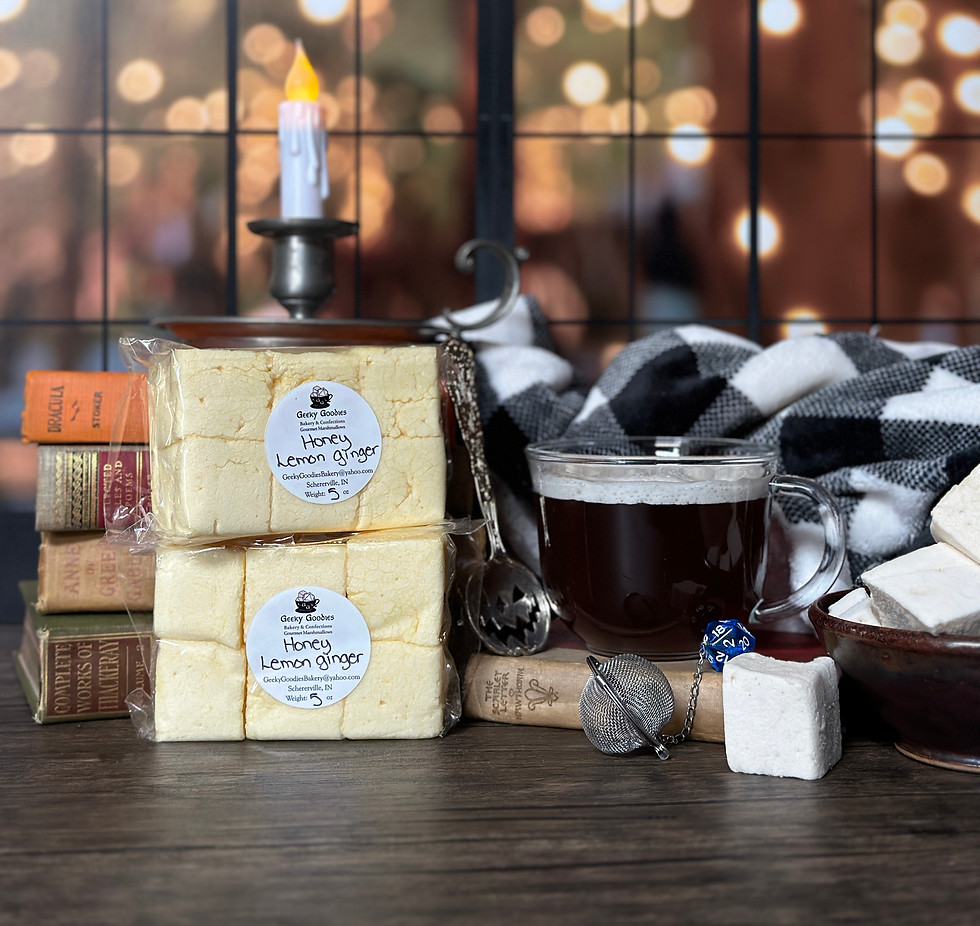 A cozy tea setup featuring two stacked packages of “Honey Lemon Ginger” marshmallows from Grey Goofer, displayed in front of a rustic window with twinkling lights. Beside them is a glass mug of dark tea, a metal tea infuser, a cork, a blue 20-sided die, and a black-and-white plaid blanket. Vintage books and a flameless candle complete the warm, inviting scene.