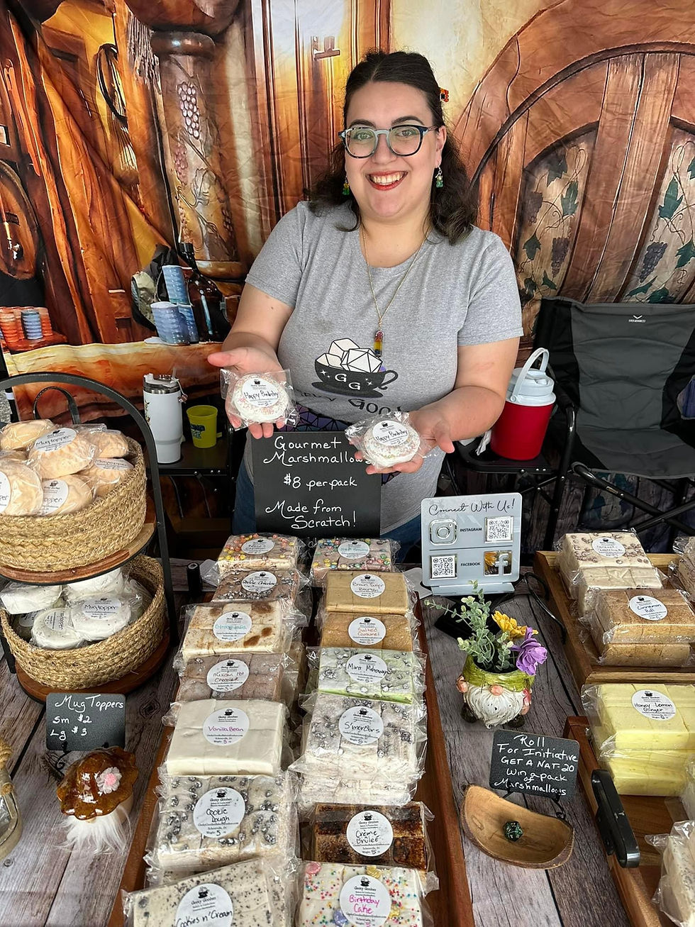 A smiling woman with glasses and long dark hair stands behind a booth displaying gourmet marshmallows. She holds up two wrapped marshmallow packs, wearing a gray t-shirt with a d20 dice graphic. The booth features handwritten signs and baskets of treats, set against a fantasy-themed backdrop resembling a wooden tavern door.