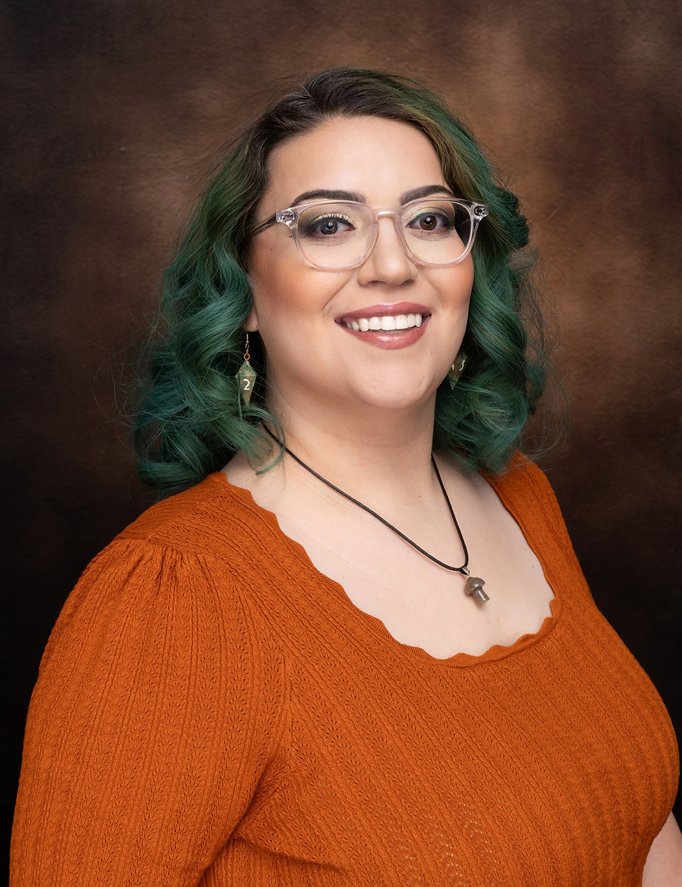 A woman with shoulder-length teal hair smiles warmly in a professional portrait. She wears clear-framed glasses, a burnt orange textured top with a scalloped neckline, and a black necklace with a small pendant. Her background is a rich brown gradient, creating a polished and inviting look.